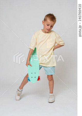 Full length studio portrait of a young boy holding a mint green penny board under his arm, looking down thoughtfully. Clean white background, perfect for youth fashion and lifestyle marketing. Active 140760410