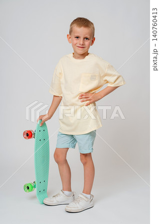 Full length portrait of a stylish young boy standing in profile, holding a vertical mint green skateboard with one hand and keeping the other on his hip. Bright minimalist studio setup. Confident 140760413
