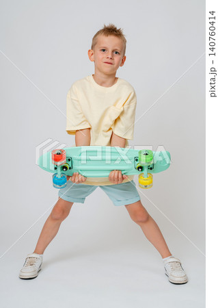 Full length portrait of a cool preteen boy holding a mint green skateboard horizontally with both hands. Confident kid posing in a bright studio for a commercial lifestyle stock photo. Active 140760414