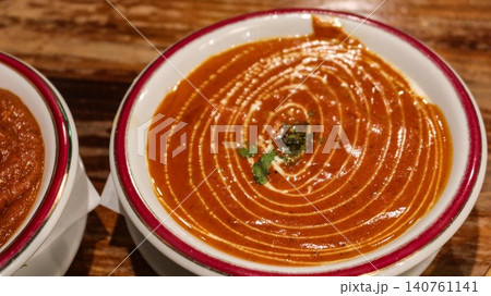 close up of Indian curry bowl with butter chicken garnished with cilantro leaves and ginger in a white bowl with red rim on wooden table with copy space 140761141