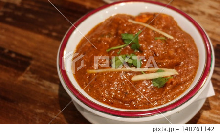 close up of Indian curry bowl with butter chicken garnished with cilantro leaves and ginger in a white bowl with red rim on wooden table with copy space 140761142