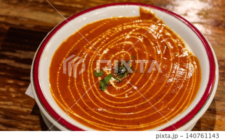 close up of Indian butter chicken curry with cream swirl and cilantro herb garnish with cream swirl on top in a white bowl with red rim on wooden table and close space 140761143
