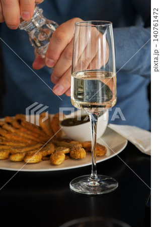 Person seasoning fried food with salt while holding a glass of white wine at a dining table Person seasoning fried food with salt while holding a glass of white wine at a dining table 140761472