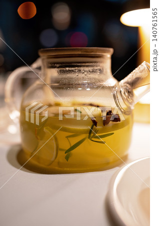 Glass teapot with herbal infusion containing lemon slices and herbs on a table with soft lighting Glass teapot with herbal infusion containing lemon slices and herbs on a table with soft lighting 140761565