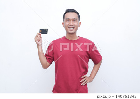 Young man smiles, holding a credit card. He wears a red t-shirt. His posture shows confidence and ease. The white background is clean and simple. Ideal for financial, shopping 140761845