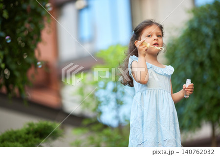 Girl in blue dress enjoying outdoor playtime, blowing soap bubbles, embodying childhood innocence and fun during summer 140762032