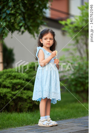 Little girl wearing a blue dress and white sandals standing on a paved path in a park holding a lollipop Little girl wearing a blue dress and white sandals standing on a paved path in a park holding a lollipop 140762046
