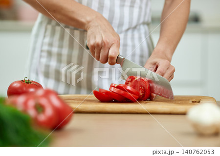 Person's hands cutting juicy red tomato slices on a wooden cutting board in a kitchen during food preparation Person's hands cutting juicy red tomato slices on a wooden cutting board in a kitchen during food preparation 140762053