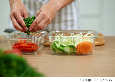 Woman's hands placing fresh parsley into a glass container with cut vegetables on a wooden kitchen table Woman's hands placing fresh parsley into a glass container with cut vegetables on a wooden kitchen table 140762055