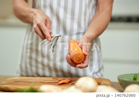 Woman's hands using a peeler to remove skin from a raw carrot, preparing fresh vegetables for cooking in a kitchen 140762060