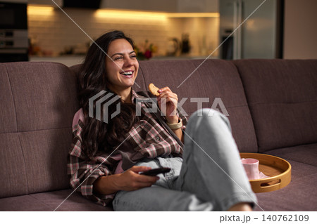 Woman feeling happy, laughing and relaxing on a sofa at home, eating a cookie and watching tv in the evening Woman feeling happy, laughing and relaxing on a sofa at home, eating a cookie and watching tv in the evening 140762109