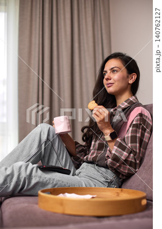 Young woman relaxing on sofa, enjoying a cookie and hot drink in her cozy living room at home. Snacking while watching tv 140762127