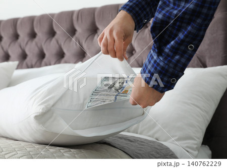 Man hiding dollar banknotes in pillow case indoors, closeup. Money savings Man hiding dollar banknotes in pillow case indoors, closeup. Money savings 140762229