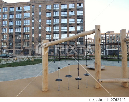 A modern, eco-style playground with wooden play structures and a rubber surface. A multi-story brick residential complex with panoramic windows is visible in the background. A modern, eco-style playground with wooden play structures and a rubber surface. A multi-story brick residential complex with panoramic windows is visible in the background. 140762442