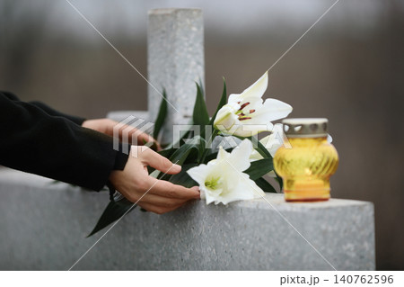 Woman holding white lilies near light grey granite tombstone with candle outdoors, closeup. Funeral ceremony Woman holding white lilies near light grey granite tombstone with candle outdoors, closeup. Funeral ceremony 140762596