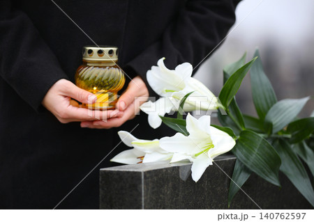Woman with candle and white lilies near black granite tombstone outdoors, closeup. Funeral ceremony Woman with candle and white lilies near black granite tombstone outdoors, closeup. Funeral ceremony 140762597
