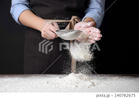 Woman sifting wheat flour at table against black background, closeup 140762675