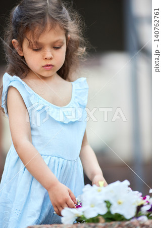 Little girl in a blue dress gently touching white and purple flowers, exploring nature and childhood innocence Little girl in a blue dress gently touching white and purple flowers, exploring nature and childhood innocence 140762761