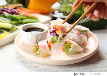 Woman eating delicious roll wrapped in rice paper at white wooden table, closeup Woman eating delicious roll wrapped in rice paper at white wooden table, closeup 140762790