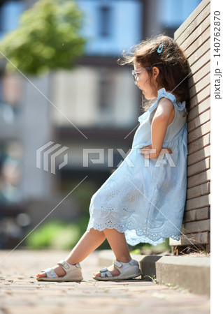 Young child in a light blue dress with glasses and an elaborate hem, leaning against a wooden wall on a summer day Young child in a light blue dress with glasses and an elaborate hem, leaning against a wooden wall on a summer day 140762870