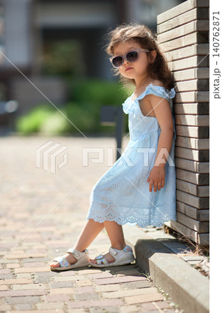 little child girl in a light blue sundress and sandals poses on a sunny urban sidewalk, playful and carefree on a warm summer day little child girl in a light blue sundress and sandals poses on a sunny urban sidewalk, playful and carefree on a warm summer day 140762871