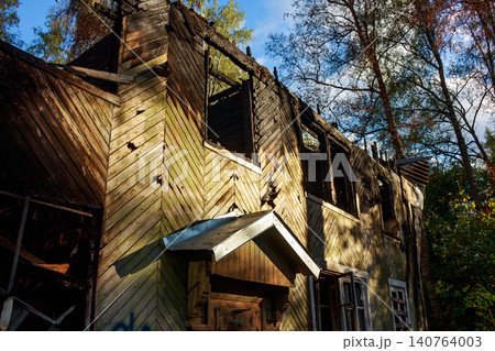A dilapidated wooden house stands partially burnt, its upper windows charred and empty, a stark image of destruction under a clear sky A dilapidated wooden house stands partially burnt, its upper windows charred and empty, a stark image of destruction under a clear sky 140764003