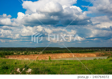 Rolling white and gray clouds dominate a vivid blue sky, overlooking a sandy quarry pond nestled in lush green fields and a distant forest Rolling white and gray clouds dominate a vivid blue sky, overlooking a sandy quarry pond nestled in lush green fields and a distant forest 140764072
