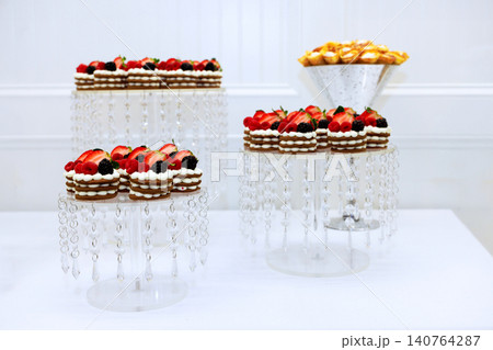 Cakes topped with berries sit on clear stands while bowl of pastries rests nearby in decorated space. Cakes topped with berries sit on clear stands while bowl of pastries rests nearby in decorated space. 140764287
