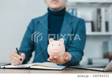 Piggy Bank and Financial Planning Concept. Businesswoman holding a pink piggy bank with a notebook and laptop on the desk, symbolizing financial planning, savings, investment, and money management. Piggy Bank and Financial Planning Concept. Businesswoman holding a pink piggy bank with a notebook and laptop on the desk, symbolizing financial planning, savings, investment, and money management. 140764331