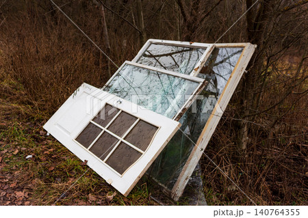 Old white window frames and a panel door with patterned glass dumped in a gloomy autumn forest, representing renovation waste and environmental pollution 140764355