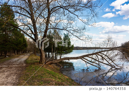 Spring flood overflowing a quiet country riverside, featuring a dramatic fallen tree stretching over tranquil waters under a bright blue sky with soft white clouds, early April seasonal landscape Spring flood overflowing a quiet country riverside, featuring a dramatic fallen tree stretching over tranquil waters under a bright blue sky with soft white clouds, early April seasonal landscape 140764358