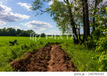 Bare firebreak carved into a lush green field, separating wild grass from dense woodland under a sunny summer sky with scattered fluffy clouds and a parked electric bike nearby Bare firebreak carved into a lush green field, separating wild grass from dense woodland under a sunny summer sky with scattered fluffy clouds and a parked electric bike nearby 140764381