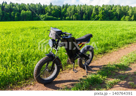 Black electric bike parked on a dirt path near a lush green field during a sunny summer day, modern eco-friendly transport for adventure and off-road riding in nature Black electric bike parked on a dirt path near a lush green field during a sunny summer day, modern eco-friendly transport for adventure and off-road riding in nature 140764391