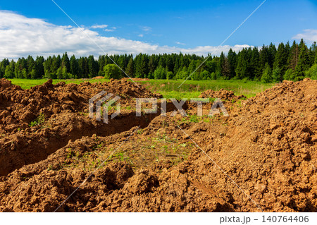 Freshly dug trench in loamy soil across an open field, showcasing raw earthworks with mounds of clay and dirt against a lush green forest backdrop under a bright blue sky Freshly dug trench in loamy soil across an open field, showcasing raw earthworks with mounds of clay and dirt against a lush green forest backdrop under a bright blue sky 140764406