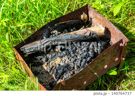 Rusty disposable metal grill left in green grass with burnt logs and charcoal ashes, an example of irresponsible camping and environmental pollution in a summer meadow setting Rusty disposable metal grill left in green grass with burnt logs and charcoal ashes, an example of irresponsible camping and environmental pollution in a summer meadow setting 140764737