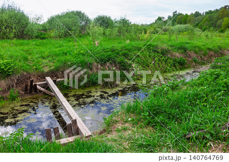 Rustic wooden plank bridge spanning a stagnant overgrown creek amidst a lush wild meadow with green bushes and young trees under a cloudy spring sky in the countryside Rustic wooden plank bridge spanning a stagnant overgrown creek amidst a lush wild meadow with green bushes and young trees under a cloudy spring sky in the countryside 140764769