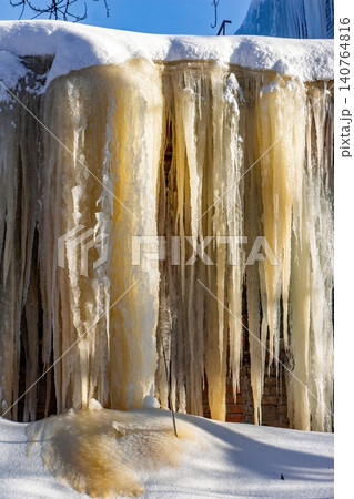 Striking golden icicles form a massive frozen curtain on a building facade, bathed in crisp morning light. A unique urban winter spectacle Striking golden icicles form a massive frozen curtain on a building facade, bathed in crisp morning light. A unique urban winter spectacle 140764816