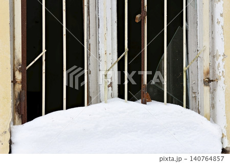 Dilapidated window with rusted bars and broken glass peeking above a mound of fresh winter snow 140764857