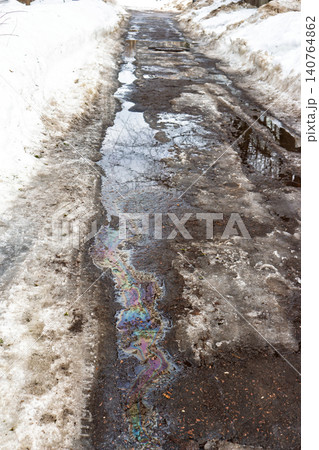 Iridescent oil sheen on a slushy spring puddle, a striking symbol of vehicle pollution on a thawing road 140764862