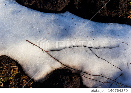 Melting snow patch on dark earth, thin branches lie across the surface, hint of early spring awakening Melting snow patch on dark earth, thin branches lie across the surface, hint of early spring awakening 140764870