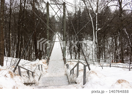 Pedestrian suspension bridge blanketed in fresh snow, spanning a serene winter forest. Gentle flakes fall, creating a quiet, picturesque scene Pedestrian suspension bridge blanketed in fresh snow, spanning a serene winter forest. Gentle flakes fall, creating a quiet, picturesque scene 140764888
