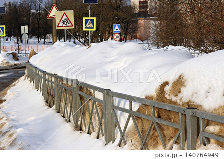 A metal roadside barrier parallels deep snowdrifts beside a road, with traffic signs visible in the background, Russia 140764949