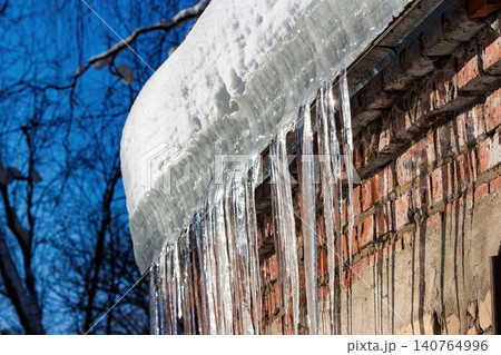 Sparkling icicles dangle from a snow-laden roof edge against a rustic brick wall. Winter's grip softens under the clear blue sky, hinting at spring's thaw Sparkling icicles dangle from a snow-laden roof edge against a rustic brick wall. Winter's grip softens under the clear blue sky, hinting at spring's thaw 140764996