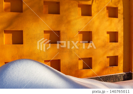 Large fluffy snowdrift rests against a vibrant orange wall featuring geometric recessed square niches under soft winter sunlight creating a striking minimalist contrast between cold and warmth Large fluffy snowdrift rests against a vibrant orange wall featuring geometric recessed square niches under soft winter sunlight creating a striking minimalist contrast between cold and warmth 140765011