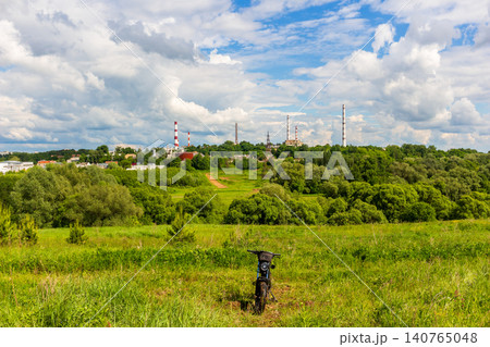 Modern electric bicycle parked in a vibrant green meadow under a cloudy blue sky with a distant industrial factory and tall smokestacks on the horizon on a sunny day Modern electric bicycle parked in a vibrant green meadow under a cloudy blue sky with a distant industrial factory and tall smokestacks on the horizon on a sunny day 140765048