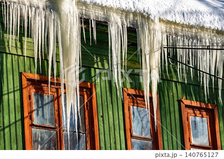Sunlit, sharp icicles pierce the winter sky, hanging from an aged green timber dwelling. Bright contrast against the weathered facade, Eastern Europe 140765127