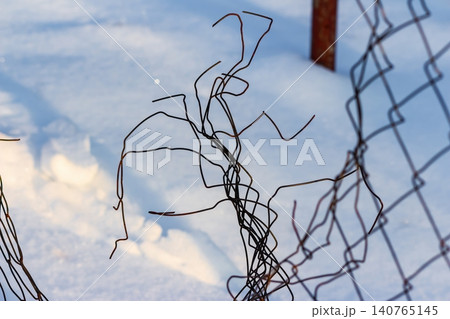 Rusted, twisted wires of a broken chain-link fence create an intricate, abstract pattern against pristine winter snow, bathed in soft evening sunlight 140765145