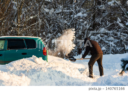 Man vigorously shoveling deep snow, clearing a path for his teal car on a bright winter day after heavy snowfall. Balkan cyclone Francis in Central Russia, January 2026 140765166
