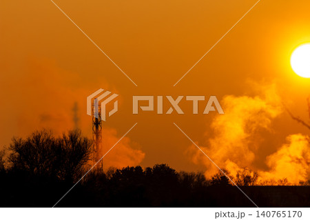 Towering industrial stack emits sunlit steam and smoke into a vivid orange sky during a breathtaking winter sunset Towering industrial stack emits sunlit steam and smoke into a vivid orange sky during a breathtaking winter sunset 140765170