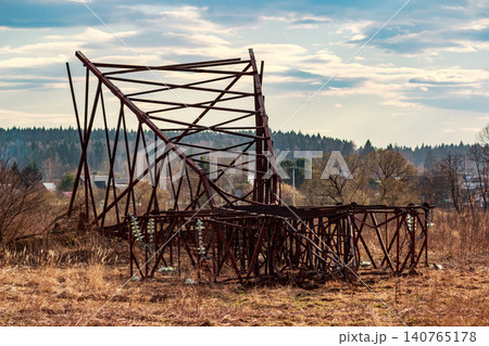 Deconstructed rusty metal electricity pylon lies fallen in a dry grassy field against a backdrop of distant trees and a cloudy spring sky Deconstructed rusty metal electricity pylon lies fallen in a dry grassy field against a backdrop of distant trees and a cloudy spring sky 140765178